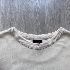 Puede incluir: Primer plano de una sudadera crema con cuello en V y ribetes acanalados. Un peque&ntilde;o logotipo rojo de Nike es visible cerca del cuello. La parte inferior de la sudadera presenta letras azul oscuro.