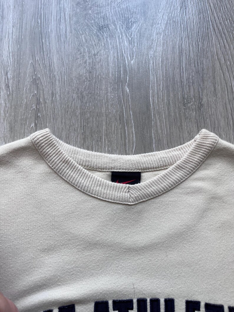 Puede incluir: Primer plano de una sudadera crema con cuello en V y ribetes acanalados. Un peque&ntilde;o logotipo rojo de Nike es visible cerca del cuello. La parte inferior de la sudadera presenta letras azul oscuro.