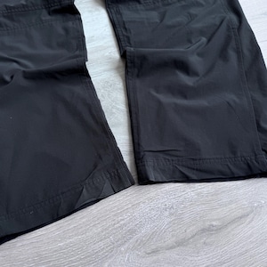 Puede incluir: Primer plano de unos pantalones negros con dise&ntilde;o de pernera recta. Los pantalones est&aacute;n hechos de un material liso, posiblemente resistente al agua. La imagen muestra la parte inferior de los pantalones, con costuras visibles y un dobladillo limpio. La tela parece tener un ligero brillo.