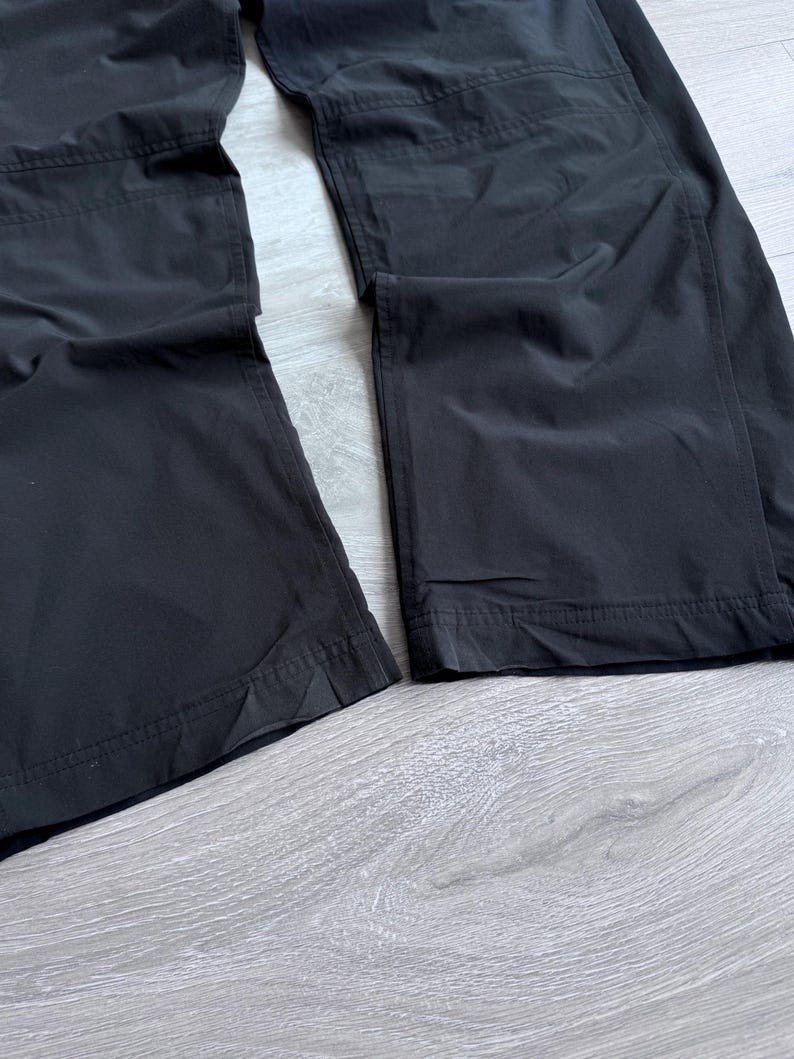 Puede incluir: Primer plano de unos pantalones negros con dise&ntilde;o de pernera recta. Los pantalones est&aacute;n hechos de un material liso, posiblemente resistente al agua. La imagen muestra la parte inferior de los pantalones, con costuras visibles y un dobladillo limpio. La tela parece tener un ligero brillo.