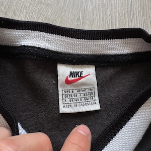 Puede incluir: Camiseta deportiva negra con cuello a rayas blancas y negras. El logotipo de Nike es visible en una etiqueta con informaci&oacute;n de talla y altura. La etiqueta tambi&eacute;n indica que la camiseta fue fabricada en Indonesia.