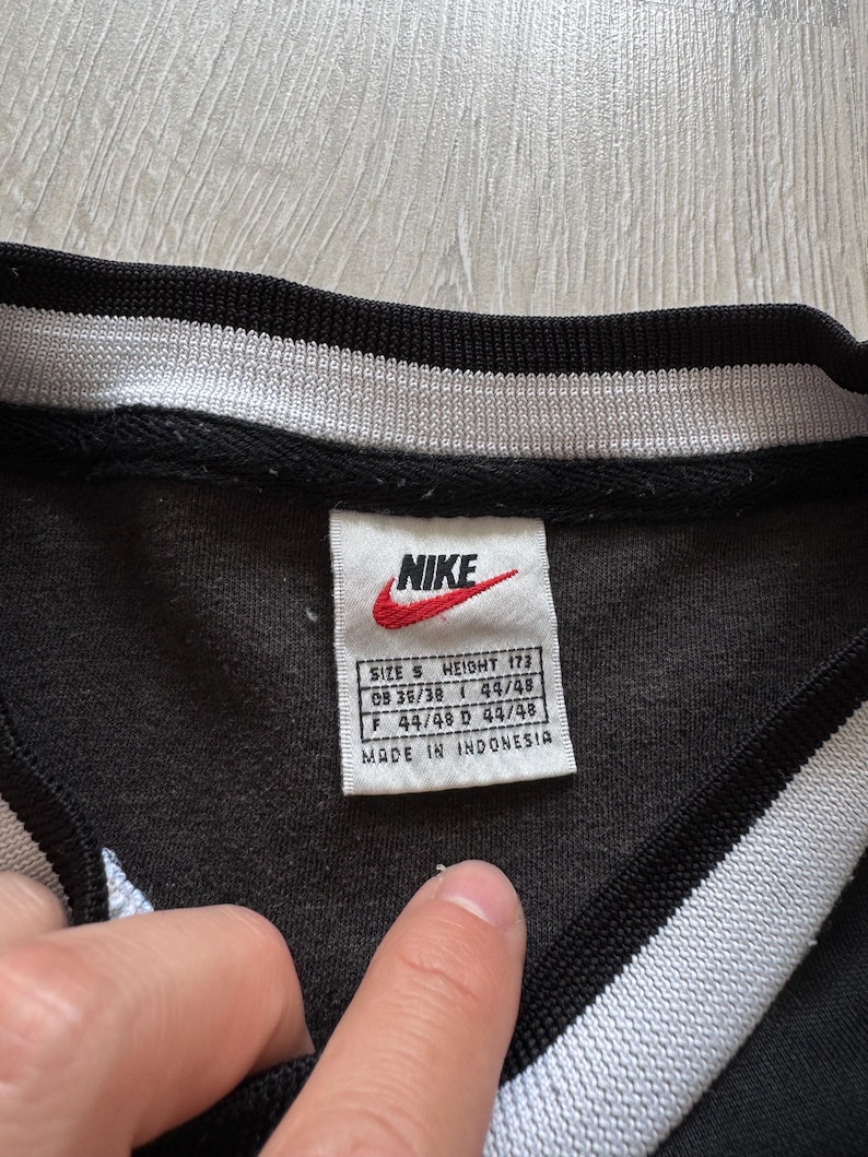 Puede incluir: Camiseta deportiva negra con cuello a rayas blancas y negras. El logotipo de Nike es visible en una etiqueta con informaci&oacute;n de talla y altura. La etiqueta tambi&eacute;n indica que la camiseta fue fabricada en Indonesia.