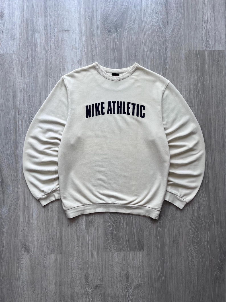 Puede incluir: Sudadera color crema con la inscripci&oacute;n "NIKE ATHLETIC" en letras negras en el pecho. La parte superior de manga larga tiene cuello redondo y pu&ntilde;os acanalados. La sudadera es de material suave.