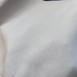 May include: Close-up of a light gray sweatshirt with a navy blue Nike swoosh logo embroidered on the upper right chest. The fabric has a textured appearance, and the bottom hem is ribbed.