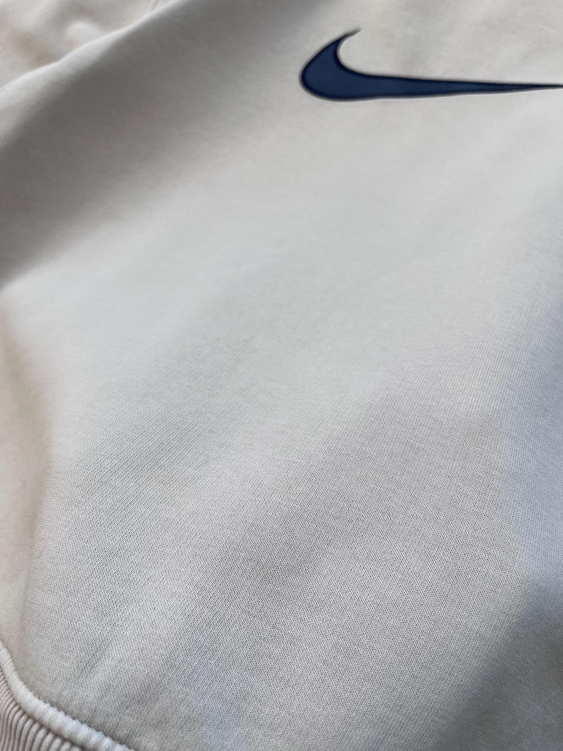 May include: Close-up of a light gray sweatshirt with a navy blue Nike swoosh logo embroidered on the upper right chest. The fabric has a textured appearance, and the bottom hem is ribbed.