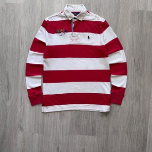 May include: A long-sleeved rugby shirt with horizontal red and white stripes. The shirt features a white collar and a small embroidered emblem on the chest. A small logo is also visible on the chest.