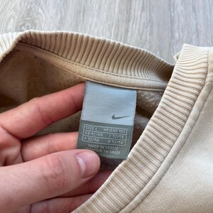 May include: Close-up of a beige Nike sweatshirt with a ribbed collar and cuffs. A size tag is visible, showing size L and height 183. The tag also includes size information for GB, F, and D. The sweatshirt is made in Greece.