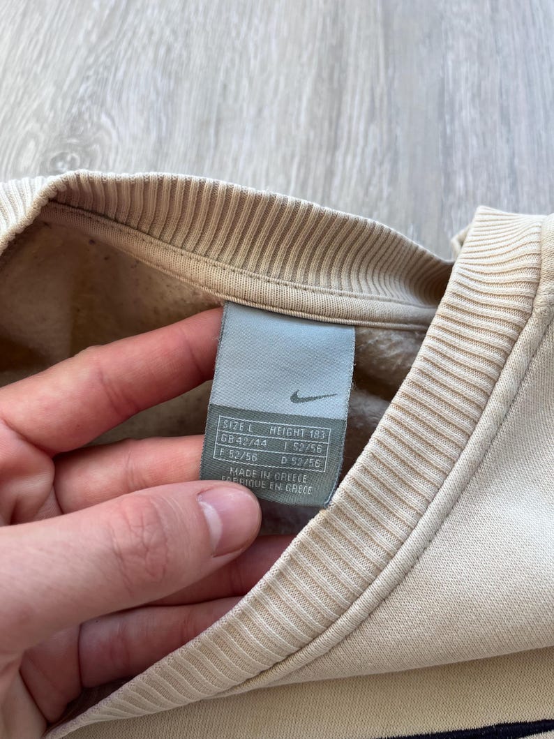 May include: Close-up of a beige Nike sweatshirt with a ribbed collar and cuffs. A size tag is visible, showing size L and height 183. The tag also includes size information for GB, F, and D. The sweatshirt is made in Greece.