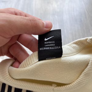 Puede incluir: Sudadera de color crema con texto negro en la parte inferior. Se ve una etiqueta negra con el logotipo de Nike e informaci&oacute;n del producto. La sudadera est&aacute; hecha de un material suave y c&oacute;modo.