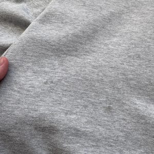 May include: Close-up of a light gray fabric, possibly a section of a garment. The fabric has a soft, textured appearance with subtle variations in tone. The image highlights the material's weave and overall color.