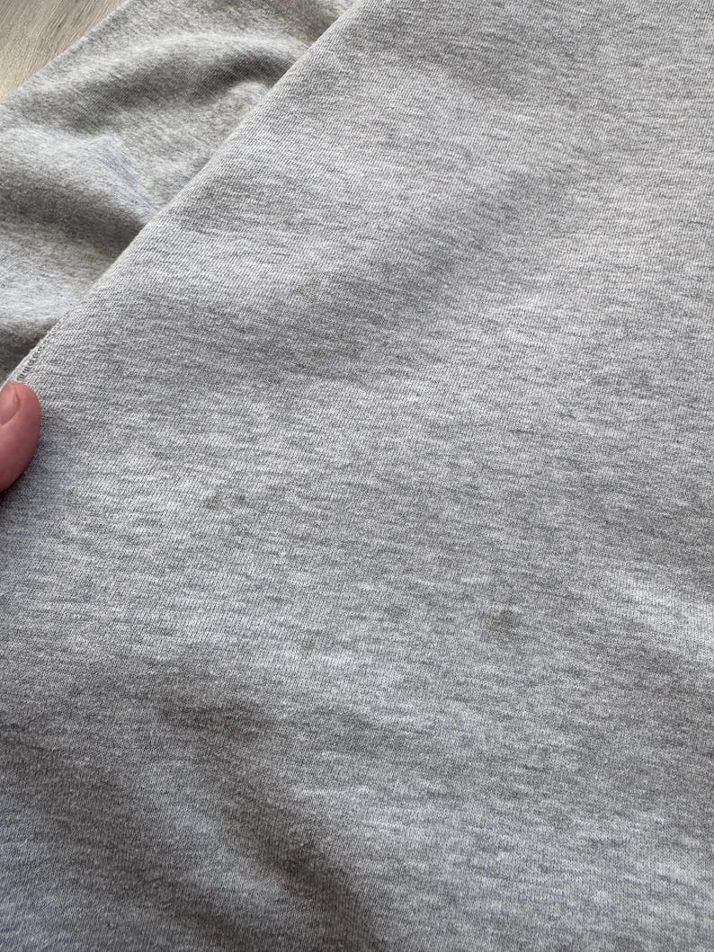 May include: Close-up of a light gray fabric, possibly a section of a garment. The fabric has a soft, textured appearance with subtle variations in tone. The image highlights the material's weave and overall color.