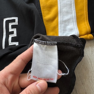 Puede incluir: Sudadera negra con un logotipo "E" blanco y rayas verticales amarillas y blancas. La etiqueta dice "KEEP AWAY FROM FIRE".