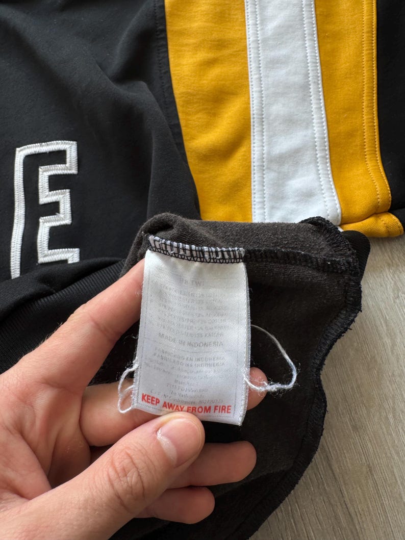 Puede incluir: Sudadera negra con un logotipo "E" blanco y rayas verticales amarillas y blancas. La etiqueta dice "KEEP AWAY FROM FIRE".