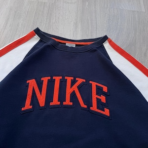 May include: Navy blue sweatshirt with the word "NIKE" in orange block letters across the chest. The sweatshirt has white and red stripes on the sleeves and a red trim around the collar.