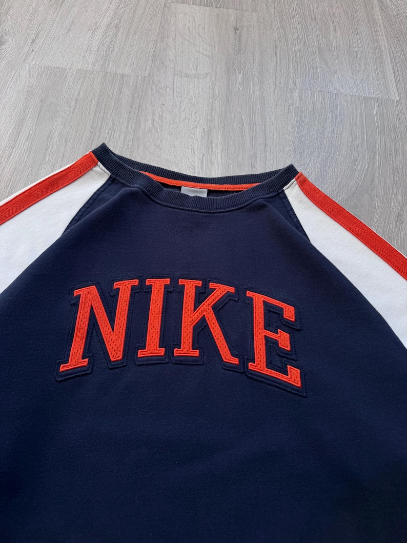 May include: Navy blue sweatshirt with the word "NIKE" in orange block letters across the chest. The sweatshirt has white and red stripes on the sleeves and a red trim around the collar.