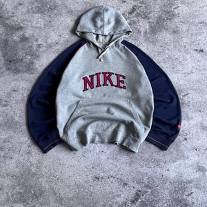 May include: A grey hooded sweatshirt featuring long navy blue sleeves and a front kangaroo pocket. The word NIKE is embroidered in red with a dark outline across the chest. The garment includes a hood with matching grey drawstrings at the neck.