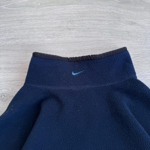 May include: A navy blue fleece pullover with a high collar and a small blue Nike logo. The collar is trimmed with black fabric. The fleece material appears soft and warm, suitable for cooler weather. The pullover is a solid color.