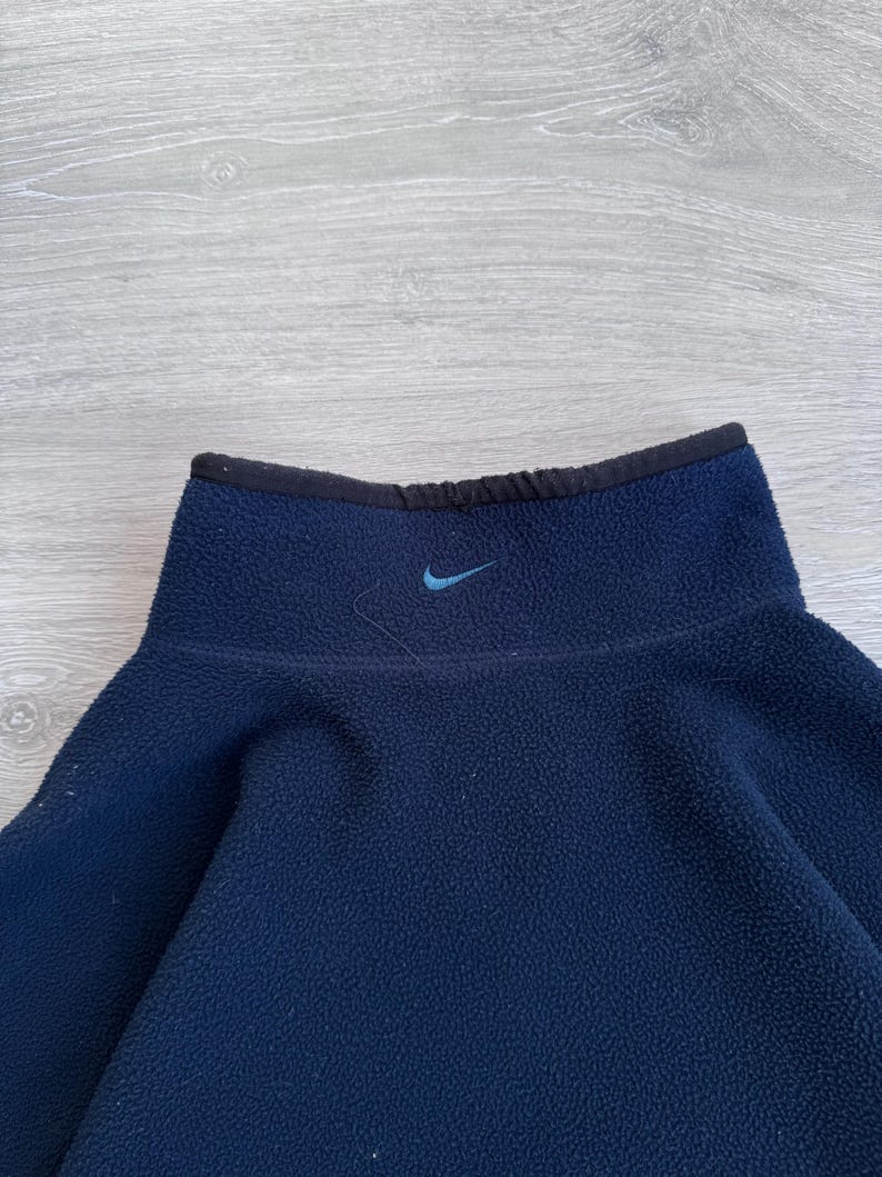 May include: A navy blue fleece pullover with a high collar and a small blue Nike logo. The collar is trimmed with black fabric. The fleece material appears soft and warm, suitable for cooler weather. The pullover is a solid color.