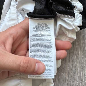 Peut inclure: Gros plan sur une &eacute;tiquette de v&ecirc;tement blanche et noire. L'&eacute;tiquette contient du texte en plusieurs langues, dont l'anglais, le fran&ccedil;ais et l'espagnol, d&eacute;taillant les mat&eacute;riaux et l'origine du produit. L'&eacute;tiquette est tenue par une main.