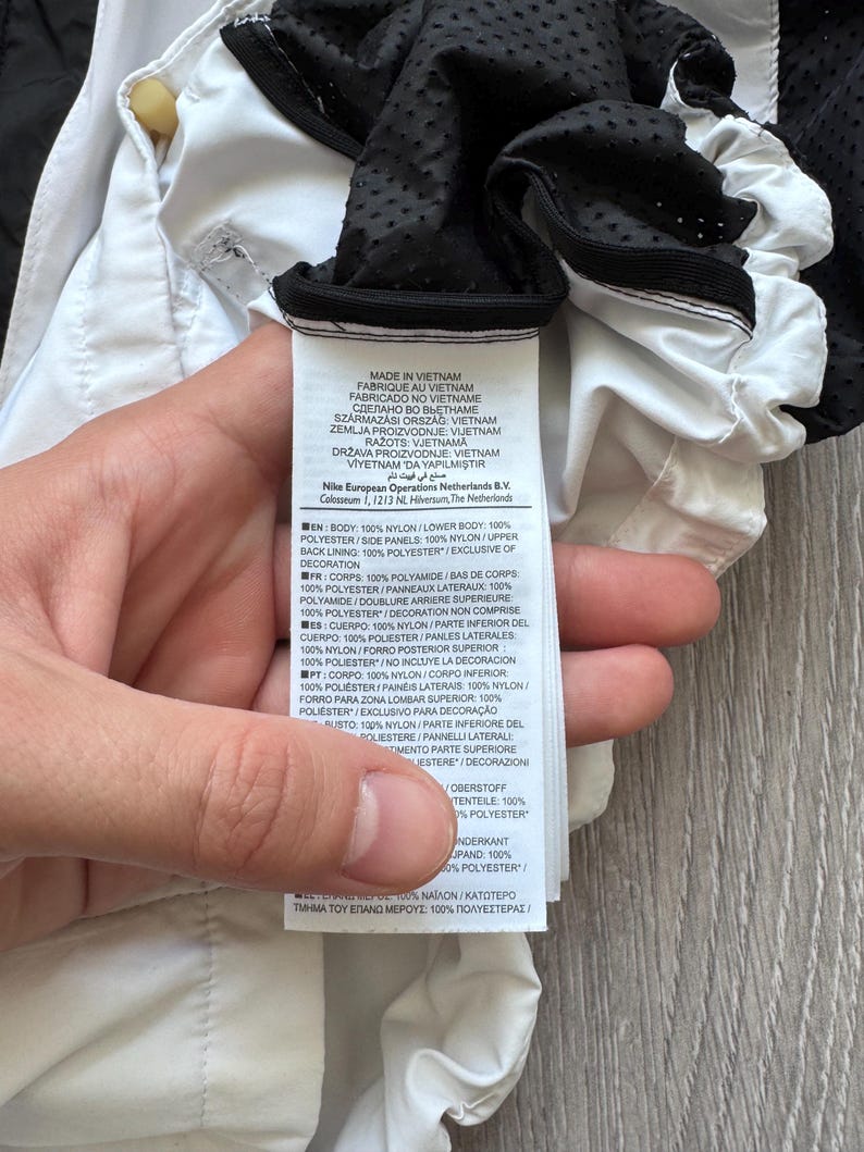 Peut inclure: Gros plan sur une &eacute;tiquette de v&ecirc;tement blanche et noire. L'&eacute;tiquette contient du texte en plusieurs langues, dont l'anglais, le fran&ccedil;ais et l'espagnol, d&eacute;taillant les mat&eacute;riaux et l'origine du produit. L'&eacute;tiquette est tenue par une main.