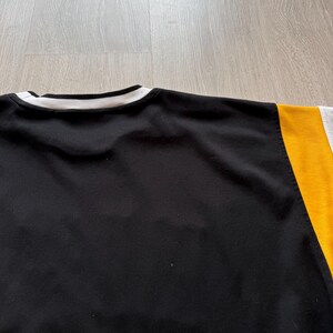 Puede incluir: Sudadera negra de cuello redondo con cuello a rayas blancas y negras. La manga derecha es amarilla y gris. La sudadera est&aacute; hecha de un material suave.