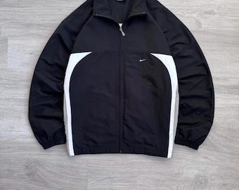 Puede incluir: Chaqueta negra con cremallera, paneles laterales blancos y un pequeño logotipo de Nike. La chaqueta tiene cuello alto y puños elásticos. La chaqueta está hecha de un material ligero.
