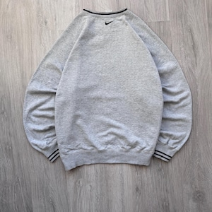 May include: A heather gray sweatshirt with a black Nike logo on the upper back. The crew neck and cuffs have black and white striped detailing. The sweatshirt is made of a soft, comfortable material.
