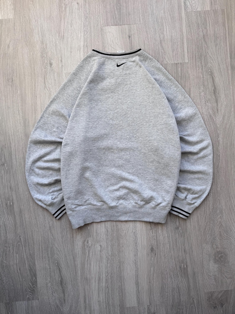 May include: A heather gray sweatshirt with a black Nike logo on the upper back. The crew neck and cuffs have black and white striped detailing. The sweatshirt is made of a soft, comfortable material.