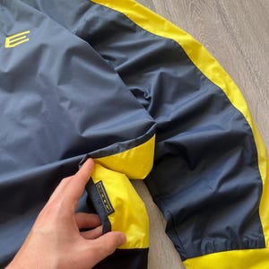May include: Navy blue and yellow athletic jacket. The jacket features a navy blue body with yellow accents along the seams and a yellow collar. The jacket has a small logo on the chest and a Nike logo on the pocket.