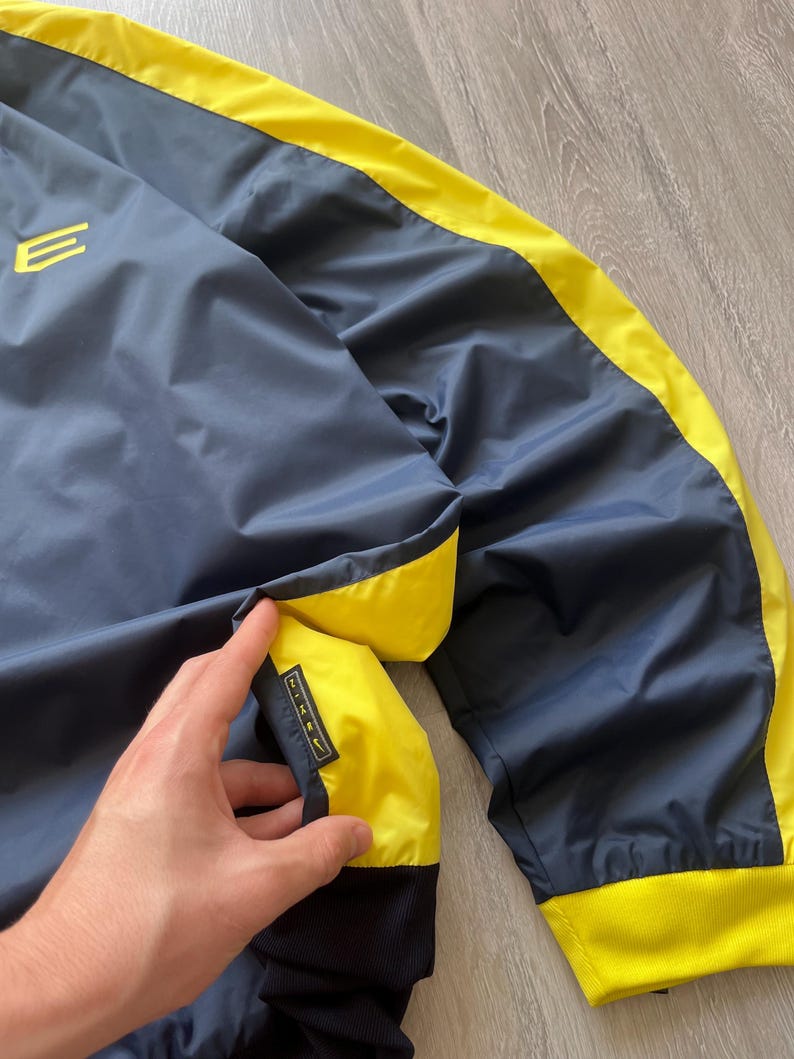 May include: Navy blue and yellow athletic jacket. The jacket features a navy blue body with yellow accents along the seams and a yellow collar. The jacket has a small logo on the chest and a Nike logo on the pocket.