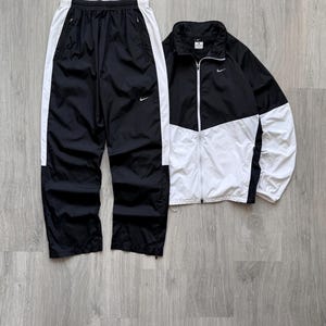 Peut inclure: Ensemble de surv&ecirc;tement Nike noir et blanc. La veste est noire et blanche avec une fermeture &eacute;clair compl&egrave;te et un petit logo Nike. Le pantalon est noir avec des rayures blanches sur les c&ocirc;t&eacute;s et un logo Nike. L'ensemble est en mati&egrave;re l&eacute;g&egrave;re.