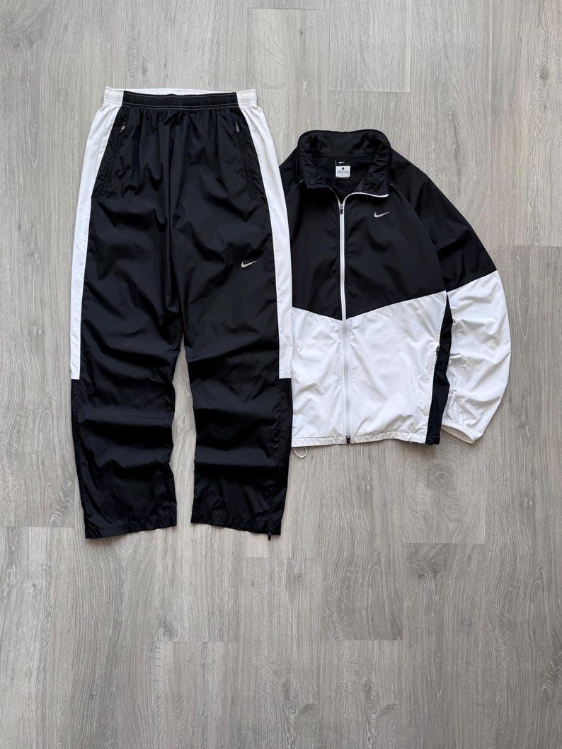 Peut inclure: Ensemble de surv&ecirc;tement Nike noir et blanc. La veste est noire et blanche avec une fermeture &eacute;clair compl&egrave;te et un petit logo Nike. Le pantalon est noir avec des rayures blanches sur les c&ocirc;t&eacute;s et un logo Nike. L'ensemble est en mati&egrave;re l&eacute;g&egrave;re.