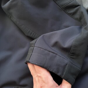 May include: Close-up of a black jacket sleeve. The sleeve is made of a smooth, matte fabric with a ribbed texture detail on the cuff. The stitching is visible along the seams, highlighting the tailored construction of the garment.