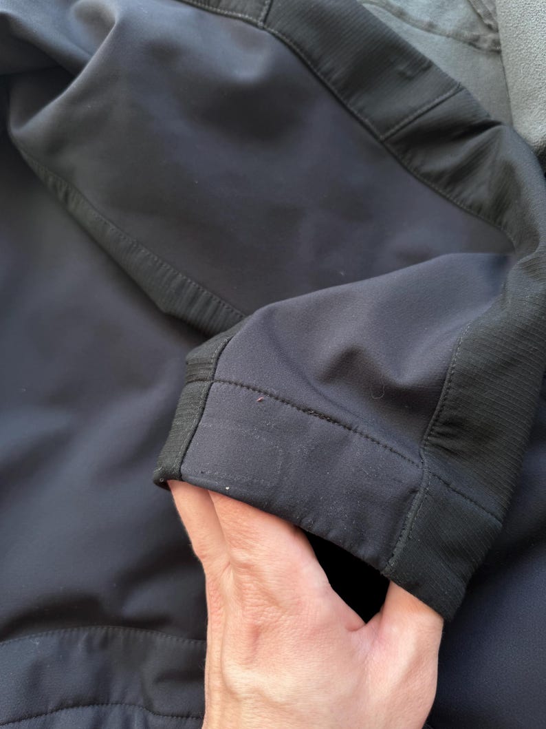 May include: Close-up of a black jacket sleeve. The sleeve is made of a smooth, matte fabric with a ribbed texture detail on the cuff. The stitching is visible along the seams, highlighting the tailored construction of the garment.