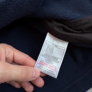 May include: Close-up of a navy blue fleece garment with a white tag. The tag has text in multiple languages, including "KEEP AWAY FROM FIRE" and "MADE IN INDONESIA". The garment is made of 100% polyester.