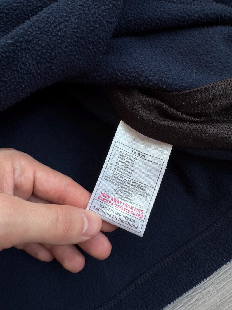 May include: Close-up of a navy blue fleece garment with a white tag. The tag has text in multiple languages, including "KEEP AWAY FROM FIRE" and "MADE IN INDONESIA". The garment is made of 100% polyester.