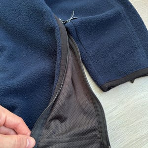May include: Navy blue fleece jacket with a full-length zipper. The jacket has a black zipper and trim. The interior lining is a dark gray mesh material. The fleece material is soft and textured.
