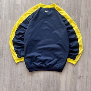 May include: A navy blue and yellow Nike windbreaker jacket. The jacket has yellow sleeves and a yellow collar, with a navy blue body. A small yellow Nike swoosh logo is visible on the upper back.
