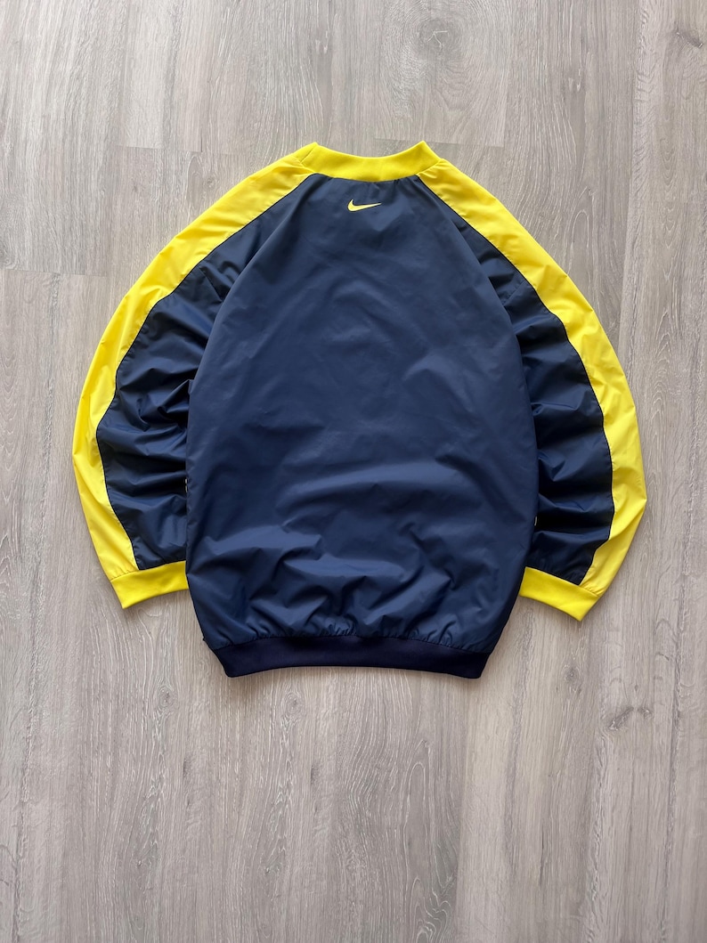 May include: A navy blue and yellow Nike windbreaker jacket. The jacket has yellow sleeves and a yellow collar, with a navy blue body. A small yellow Nike swoosh logo is visible on the upper back.