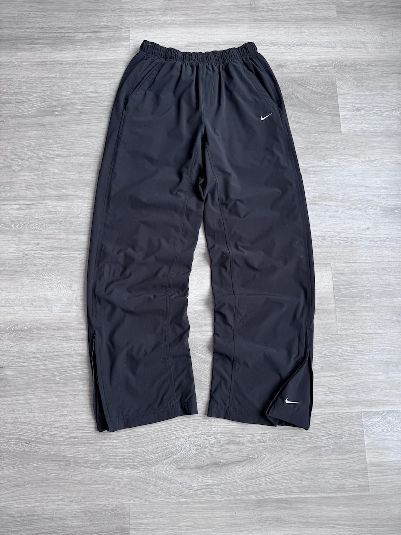 Puede incluir: Pantalones deportivos negros con un logotipo blanco de Nike en la parte delantera y cerca del tobillo. Los pantalones tienen cintura el&aacute;stica y bolsillos laterales. Las piernas tienen cremalleras en la parte inferior. Los pantalones son de material ligero.