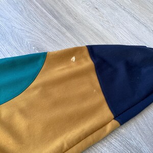 May include: Close-up of a section of a color-blocked garment. The garment features panels of teal, mustard yellow, and navy blue. The fabric appears to be a soft, possibly knit material. There are a few small spots on the mustard yellow panel.