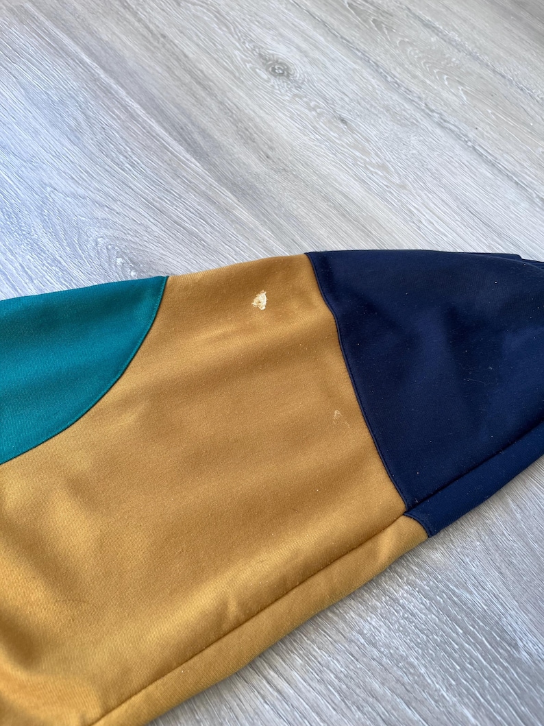 May include: Close-up of a section of a color-blocked garment. The garment features panels of teal, mustard yellow, and navy blue. The fabric appears to be a soft, possibly knit material. There are a few small spots on the mustard yellow panel.