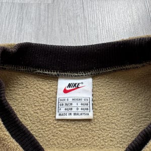 May include: Close-up of a tan and brown Nike sweatshirt. The collar and trim are dark brown. A white tag with the Nike logo and size information is visible. The tag reads "MADE IN MALAYSIA."