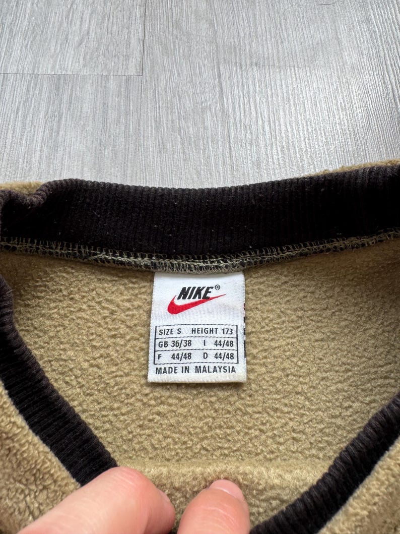 May include: Close-up of a tan and brown Nike sweatshirt. The collar and trim are dark brown. A white tag with the Nike logo and size information is visible. The tag reads "MADE IN MALAYSIA."