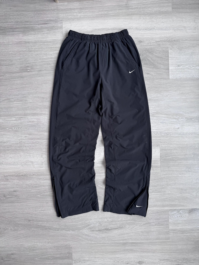Puede incluir: Pantalones de ch&aacute;ndal Nike negros con cintura el&aacute;stica y bolsillos laterales. Los pantalones presentan un peque&ntilde;o logotipo blanco de Nike en la pierna izquierda y cremalleras en los tobillos. Fabricados con un material ligero.