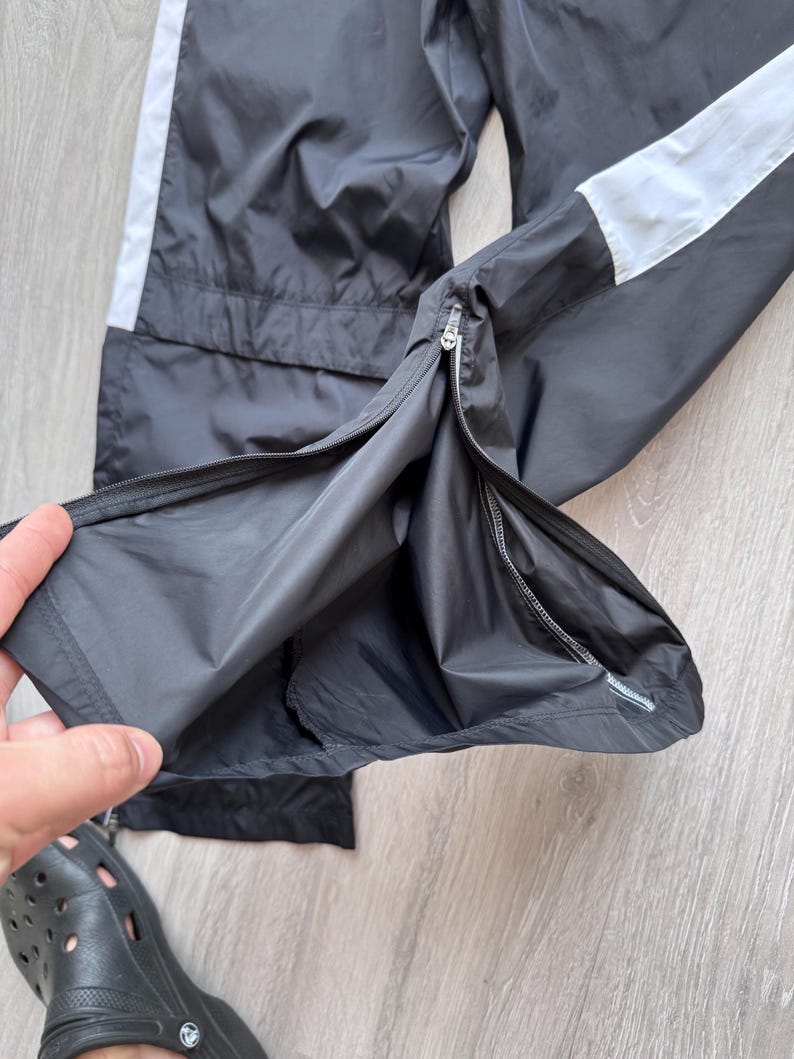 Peut inclure: Pantalon de sport noir avec une bande blanche sur le c&ocirc;t&eacute;. Le pantalon a une fermeture &eacute;clair &agrave; la cheville et est fait d'un mat&eacute;riau l&eacute;ger. Le pantalon est montr&eacute; partiellement ouvert, r&eacute;v&eacute;lant la doublure int&eacute;rieure.