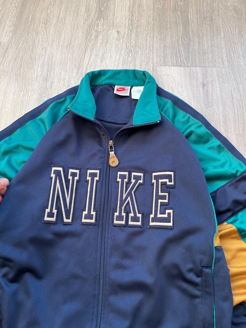 May include: Navy blue Nike track jacket with teal and gold accents. The jacket features a full zipper, a teal collar, and the Nike logo in white block letters across the chest. The jacket has a small red Nike logo on the upper chest.
