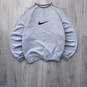 May include: A light gray sweatshirt with a black Nike swoosh logo on the chest. The crew neck and cuffs have black and white striped detailing. The sweatshirt is made of a soft, comfortable material.