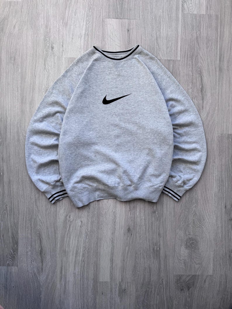 May include: A light gray sweatshirt with a black Nike swoosh logo on the chest. The crew neck and cuffs have black and white striped detailing. The sweatshirt is made of a soft, comfortable material.