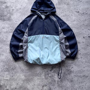 May include: A navy blue, light blue, and grey hooded windbreaker jacket featuring a half-zip closure, long sleeves, and a small gold Nike swoosh logo centered on the chest. The jacket includes two zippered side pockets and a drawstring hem.